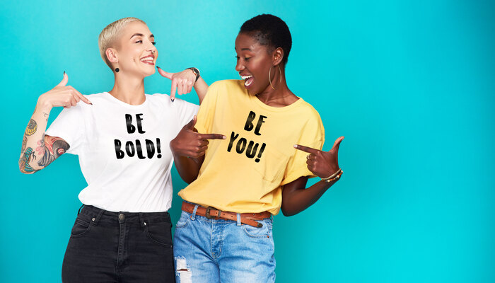 Zwei lachende Frauen mit Aufdruck auf ihren T-Shirts, Einmal Aufdruck BE BOLD! und einmal Aufdruck BE YOU!